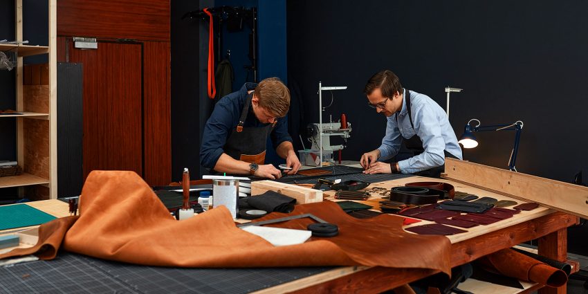 Two craftsmen working with leather and industrial tools on a large workshop table